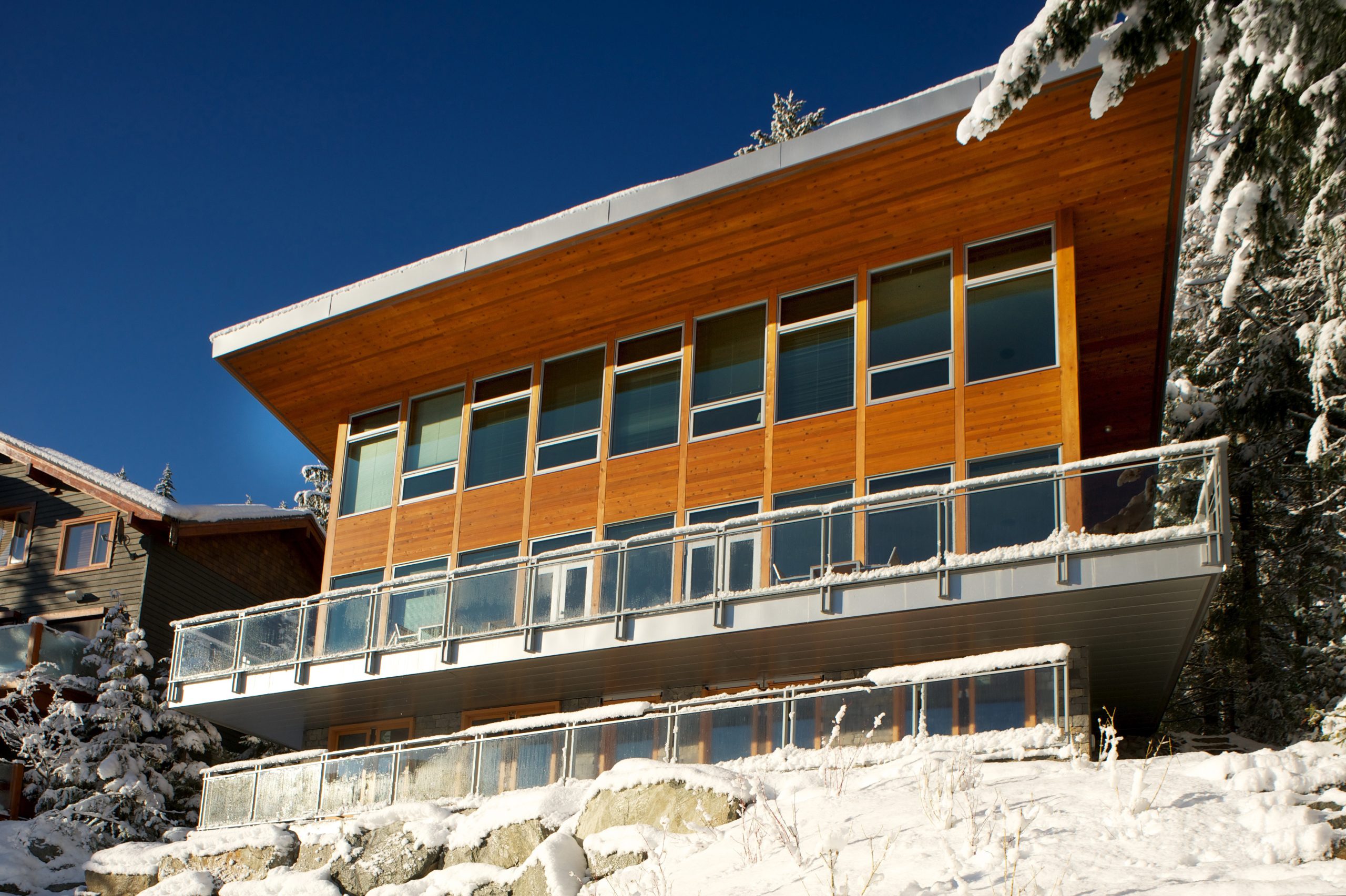 Charming wooden cabin with large windows in a snowy forest setting, surrounded by frosted trees and rocks.