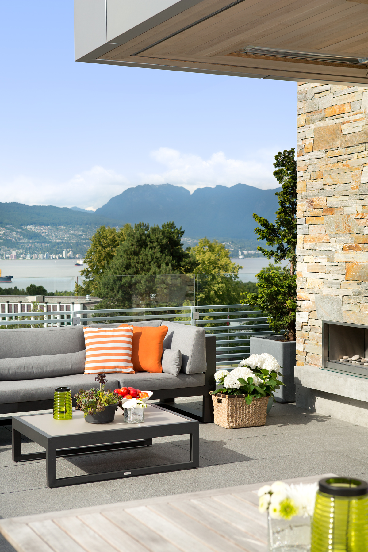 Patio designed by a top construction company, featuring a grey sofa, colorful pillows, coffee table, and stunning mountain view in the background showcasing Vancouver.