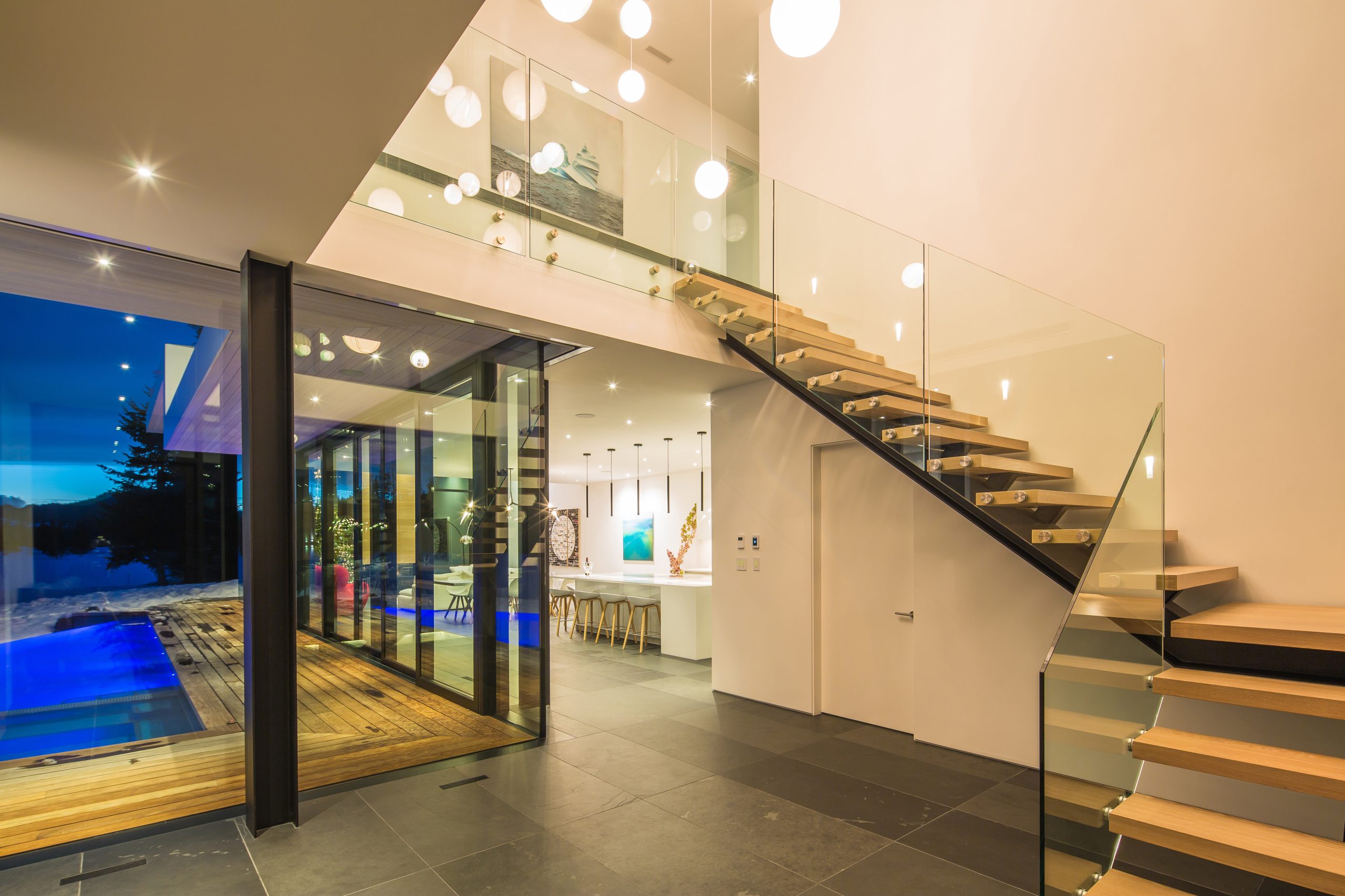 Interior with floating glass staircase, pendant lights, and view of pool through large windows at dusk, showcasing refined British Columbia construction.