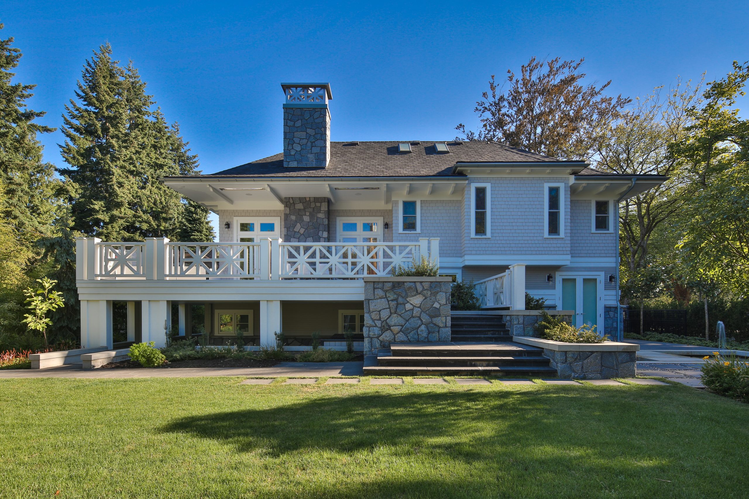 White two-story house with large porch, stone chimney, and spacious green lawn on a sunny British Columbia day.