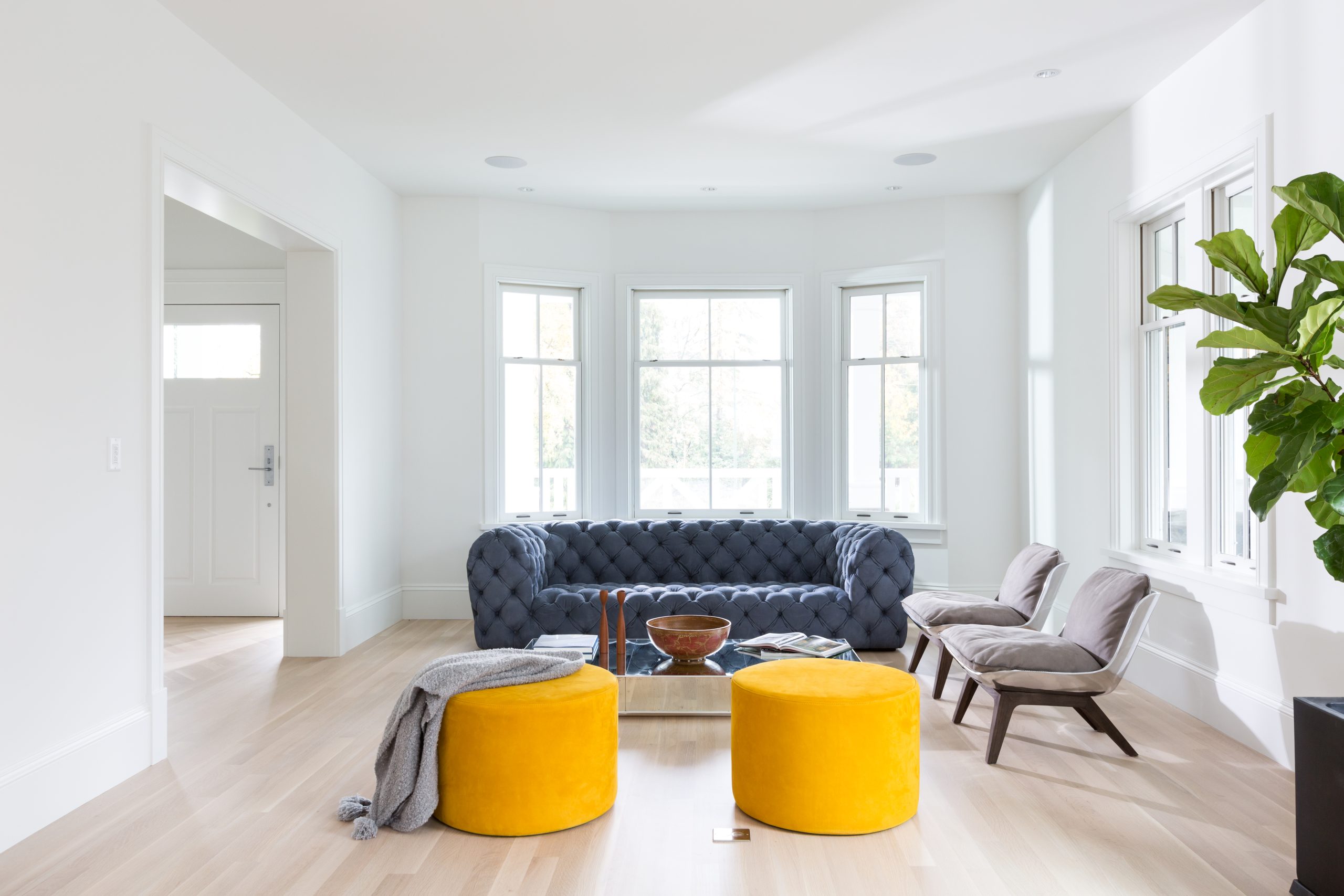 Bright living room in one of Vancouver’s quality custom homes, featuring a blue sofa, two yellow ottomans, chairs, and large windows letting in natural light.